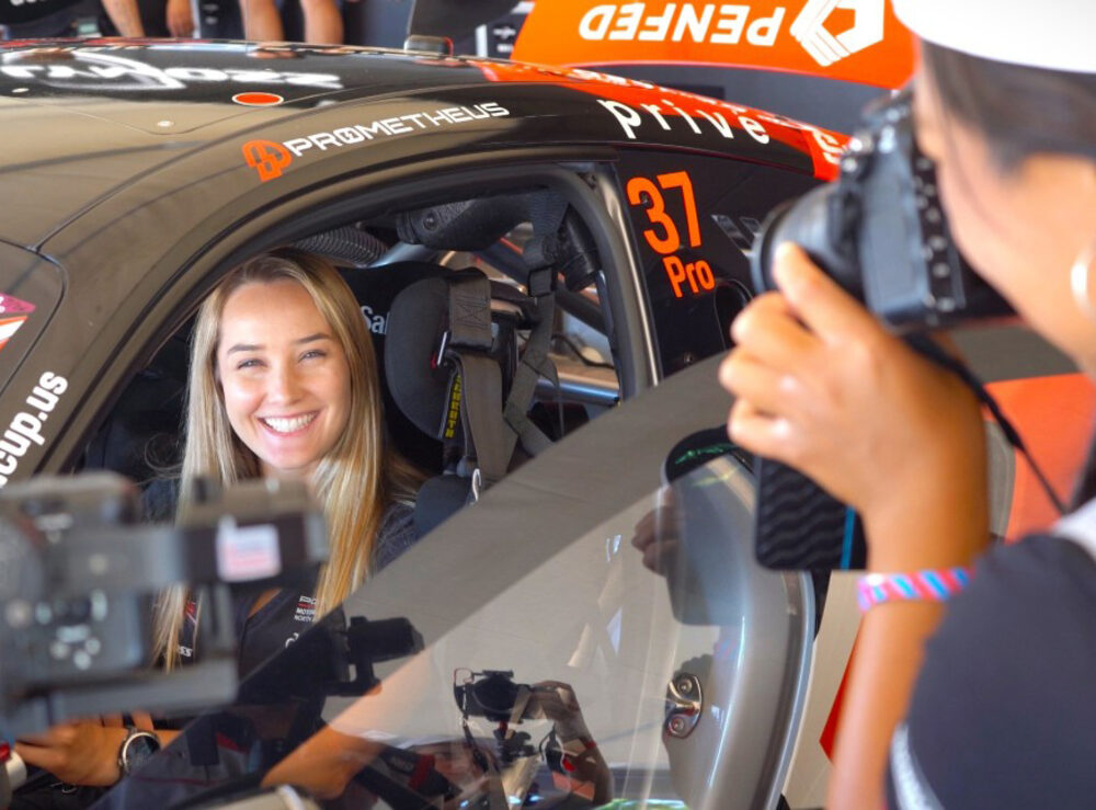 A driver in the cockpit of a race car being photographed in the paddock